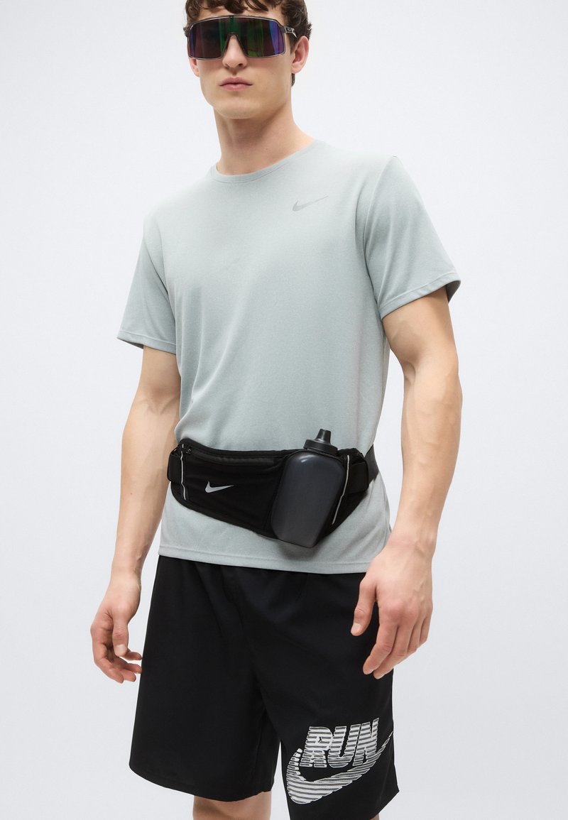 Hellgraues Nike-T-Shirt mit kurzen Ärmeln, schwarze Shorts mit "RUN"-Grafik, schwarze Bauchtasche mit Wasserflasche und bunte Sonnenbrille.