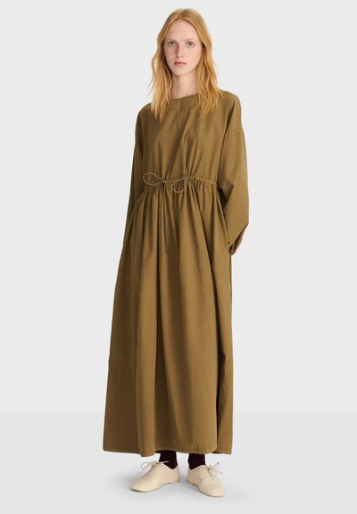 Jeune femme aux longs cheveux roux portant une robe ample olive longue jusqu'aux chevilles avec taille à cordon et chaussures blanches à lacets.
