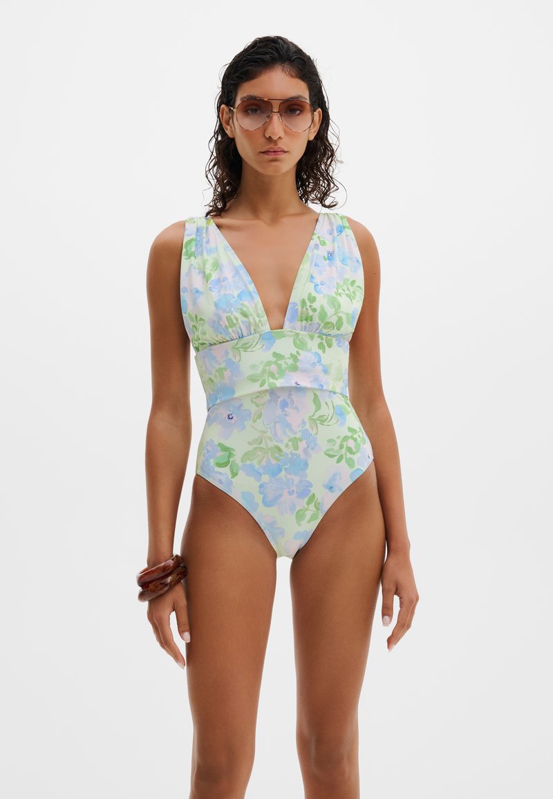 Γυναίκα που φοράει πράσινο ολόσωμο μαγιό με floral σχέδιο και βαθύ V, καφέ γυαλιά ηλίου και δύο χοντρές καφέ βραχιόλια στον αριστερό καρπό, στέκεται μπροστά σε λευκό φόντο.