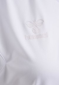 Biała tkanina jersey o subtelnej teksturze z wypukłym, jasnym logo Hummel oraz grafiką pszczółki umieszczoną centralnie.