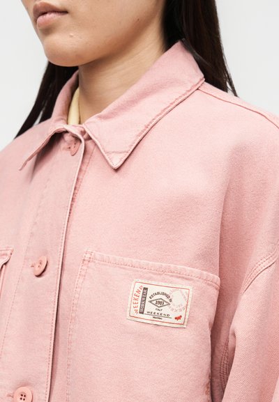 Κοντινή λήψη ενός ατόμου που φοράει ροζ πουκάμισο-μπλούζα με κουμπιά, με τσέπη στο στήθος και ένα λευκό ορθογώνιο ετικετάκι.