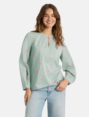 Junge Frau lächelt, trägt eine grün-weiß gestreifte Bluse mit pinken Blumenmustern und hellblauen Jeans, steht mit einer Hand in der Tasche.