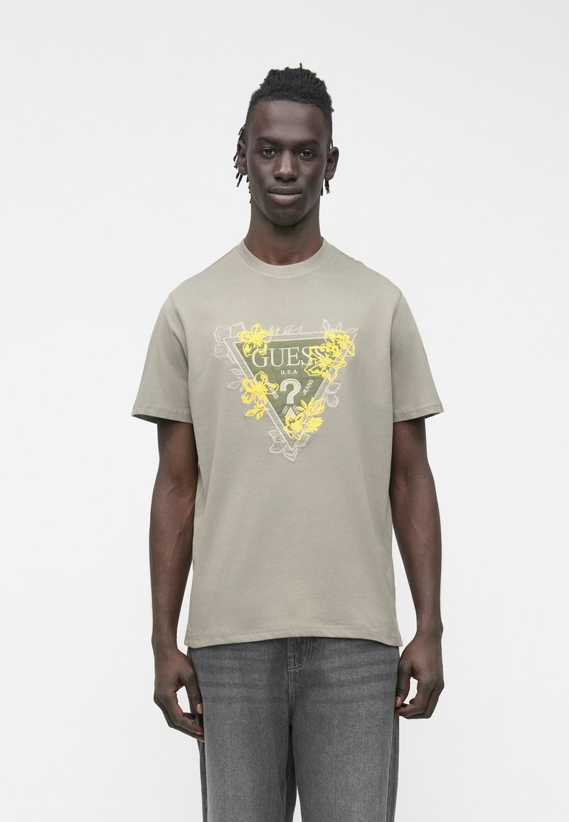 Gaiši pelēka kokvilnas T-krekls ar ziedu grafiku dzeltenā un zaļā krāsā, apaļu izgriezumu un īsām piedurknēm, kopā ar pel�ēnām džinsa biksēm.