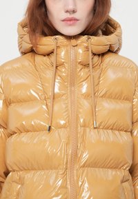 Giovane donna che indossa un giubbotto imbottito lucido color beige con cappuccio e coulisse, mostrata dalla spalla alle labbra.
