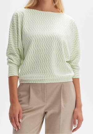 Frau trägt hellgrünes, langärmeliges Oberteil mit wellenförmigem Muster und beige, maßgeschneiderte Hosen, steht mit entspannten Armen seitlich.