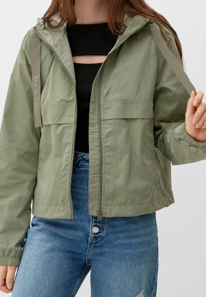 Personne portant une veste à capuche zippée vert clair sur un haut noir avec découpe, associée à un jean bleu taille haute, tenant le cordon de la veste.