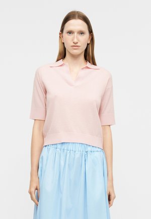 Femme aux cheveux châtain clair lisses portant un pull à manches courtes col polo de couleur rose pâle et une jupe bleue claire taille haute.