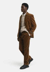 Un costume en velours côtelé marron avec un pantalon assorti, un pull en tricot beige texturé en dessous et des chaussures noires, présentant un design de revers structuré.