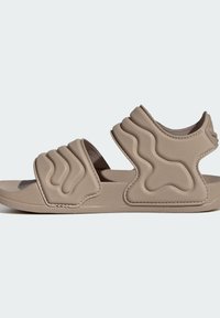 Sandale beige à bout ouvert avec des brides rembourrées texturées ondulées sur le dessus du pied et autour du talon, sur une semelle plate, sur un fond uni.