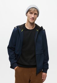 Marineblå jakke med hætte, lynlåslommer og grønne detaljer. Båret over en sort skjorte og parret med brune cargo-bukser. Grå beanie-hat.