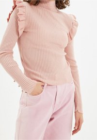 Maglione rosa a costine con spalline a volant, maniche lunghe e bottoni decorativi ai polsini. Indossato con pantaloni rosa chiaro.