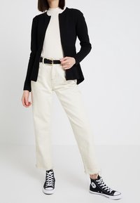 Czarna kurtka na zamek z dopasowanym fasonem, noszona na kremowej prążkowanej bluzce, w połączeniu z kremowymi, postrzępionymi dżinsami i czarnymi wysokimi trampkami.