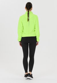 Fluorescerande grön vindjacka med hög krage och reflexdetalj på baksidan, tillsammans med svarta leggings och sneakers. Glatt, lätt material.