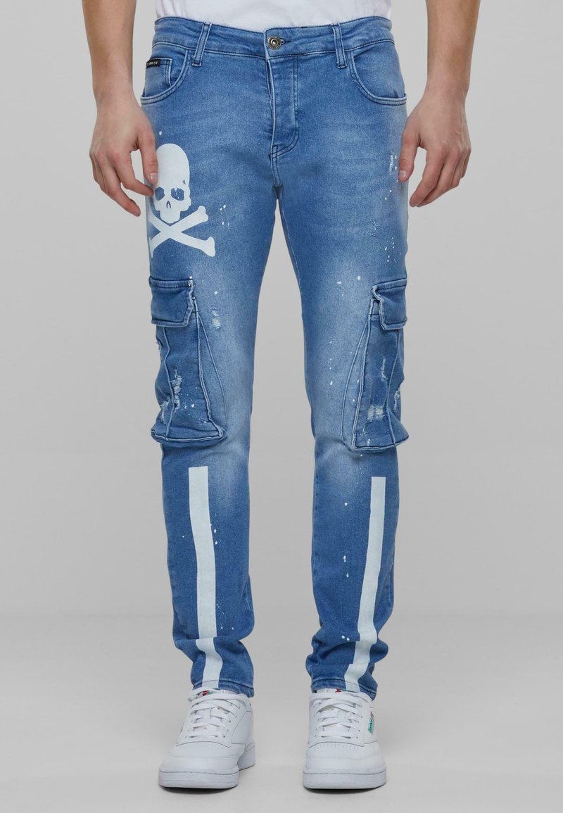 Pantalones cargo de mezclilla en azul claro con un gran gráfico de calavera y huesos cruzados en blanco, detalles de salpicaduras de pintura y franjas laterales blancas.