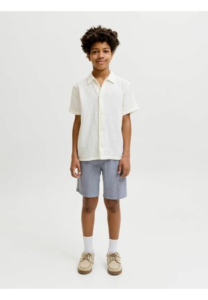 Niño con cabello rizado, de pie, lleva una camiseta blanca de manga corta, pantalones cortos grises, calcetines blancos y zapatos beige sobre un fondo liso.