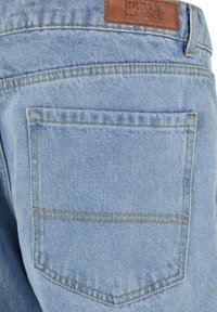 Detail zadního kapsy světlých modrých džín s oranžovým šitím a hnědým koženým štítkem s nápisem "Urban Classics Denim Factory."