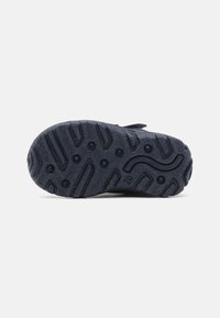 Semelle en caoutchouc noir avec surface texturée et rainures de traction, présentant un logo et de petits détails circulaires pour une meilleure adhérence.
