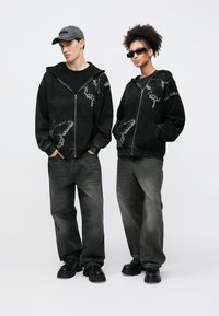 Μαύρο φούτερ με φερμουάρ και σχέδιο πεταλούδας, συνδυασμένο με χαλαρά μαύρα τζιν. Αξεσουάρ περιλαμβάνουν μαύρο καπέλο και γυαλιά ηλίου. Υφασμα με υφή.