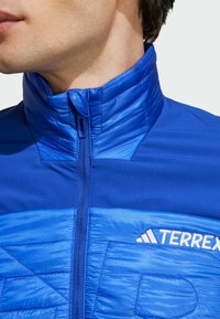 Blauwe ritsjack met hoge kraag, met gemengde texturen in de stof, een diagonaal patroon en het "TERREX"-logo in het wit.