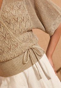 Dzianinowy top o luźnym, otwartym wzorze w kolorze beżowym, z ribbowanym dołem i związanym w talii. Teksturowany materiał dodaje głębi; rękawy są szerokie.