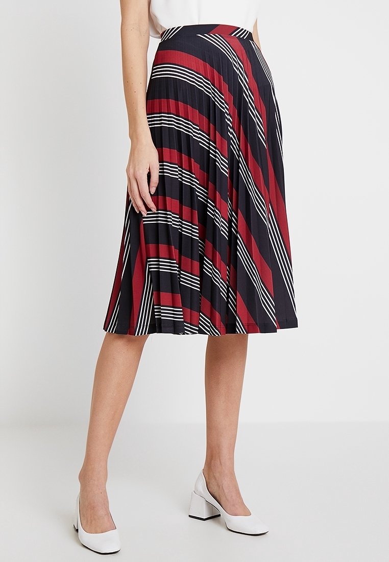 Femme portant une jupe plissée mi-longue aux genoux avec des rayures diagonales rouges, noires et blanches, associée à des chaussures blanches à talons carrés.