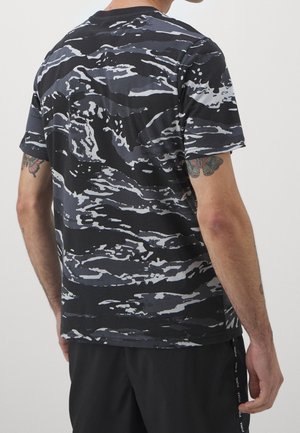 Mand iført en sort og grå t-shirt med camouflage mønster med synlige tatoveringer på begge arme, stående mod en ensfarvet baggrund.