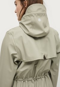 Lichtbeige waterdichte jas met een hoge kraag en een verstelbare capuchon. Heeft een elastische taille en een klein wit logo op de achterkant.