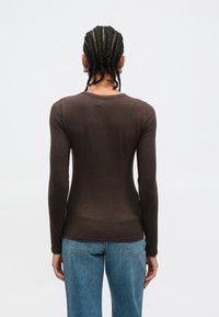 Καφέ πουκάμισο με μακριά μανίκια και στρογγυλή λαιμόκοψη, κατασκευασμένο από ελαφρύ ύφασμα, εφαρμοστός σχεδιασμός και ομαλή υφή· φορεμένο με μπλε τζιν.