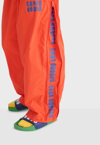 Pantalon orange vif avec le texte bleu « BOBO CHOSES » le long du côté, personne portant des chaussettes jaunes avec des sandales-claquettes bleues et vertes.