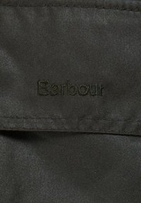 Tessuto verde scuro con una texture liscia, caratterizzato da un logo "Barbour" ricamato con filo coordinato. Dettaglio di orlo cucito visibile.