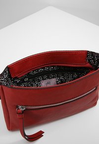 Czerwona skórzana kopertówka z zamknięciem na zamek, teksturowaną powierzchnią oraz czarnym wnętrzem w kwiatowy wzór. Wyposażona w przednią kieszeń i pasek na nadgarstek.