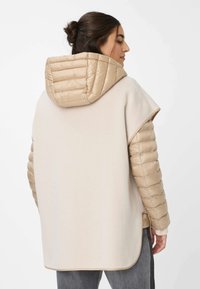 Beige oversized jas met een gewatteerde capuchon en mouwen. Textuurstof contrasteert met glad, glanzend puffermateriaal. Zijsplitten voor beweging.