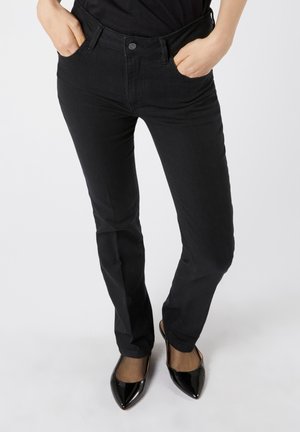 Kvinde iført smalle sorte bukser og sorte spidse højhælede sko, stående med hænderne i lommerne på en hvid baggrund.