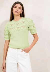 Pulover tricotat de culoare verde deschis, cu mâneci bufante, având un model din dantelă în jurul gâtului și al părții superioare a corpului. Asortat cu pantaloni albi.