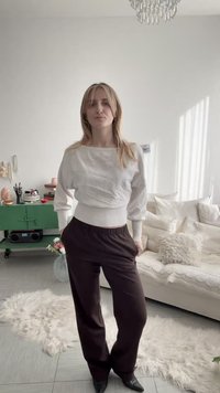 Γυναίκα με μακριά ξανθά μαλλιά στέκεται σε ένα φωτεινό σαλόνι, φορώντας λευκό πουλόβερ, μαύρο παντελόνι και μαύρα μυτερά παπούτσια, με τα χέρια στις τσέπες.