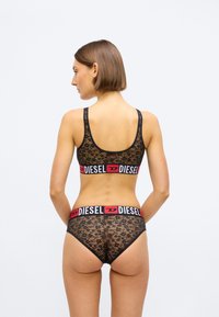 Μαύρο δαντελένιο μπραλέτ και αντίστοιχο σλιπ, με πλατιές ελαστικές ζώνες σε κόκκινο και μαύρο χρώμα με επωνυμία, διακοσμημένα με λουλουδάτο μοτίβο και αόρατες άκρες.