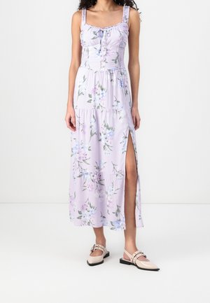 Kwiatowa sukienka maxi w jasnym fioletowym kolorze z marszczonymi ramiączkami na ramionach, guzikami z przodu oraz rozcięciem na udzie. Noszona z jasnym obuwiem.