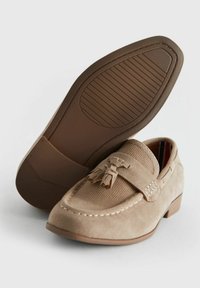 Mocassini beige in suede con tomaia testurizzata, dettaglio con nappine e tacco basso impilato. La suola in gomma presenta scanalature sottili per una migliore aderenza.