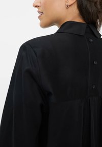 Maglietta nera con colletto classico, chiusura a bottoni sul retro e dettagli plissé. Il tessuto appare liscio e leggero.