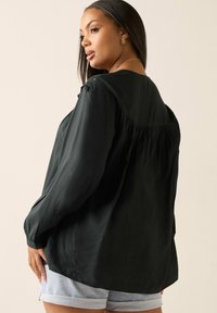 Czarna bluzka z długim rękawem o luźnym kroju, z plisami z tyłu i guziczkami na ramionach, zestawiona z jasnymi dżinsowymi szortami.
