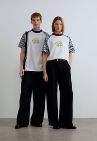 Λευκά T-shirts με μανίκια σε μαύρο ζιγκ-ζαγκ σχέδιο και κίτρινο λογότυπο, σε συνδυασμό με φαρδιά μαύρη παντελόνια. Ομαλό ύφασμα, άνετη εφαρμογή.