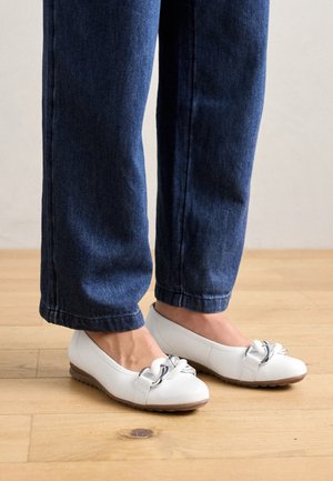 Fødder iført hvide balletsko med dekorative sølvknuder, kombineret med mørkeblå jeans med lige ben på et lyst trægulv.