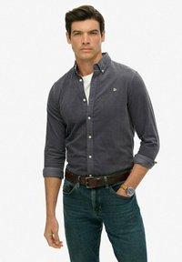 Man in een donkergrijs overhemd met knopen, witte onderhemd, blauwe jeans, een leren riem en een horloge, met één hand in zijn zak.