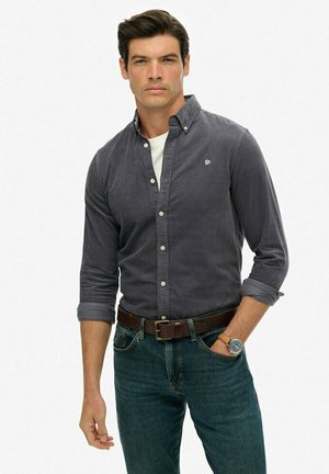 Man wearing dark gray button-up shirt, white undershirt, blue jeans, brown belt, and wristwatch, with one hand in pocket.