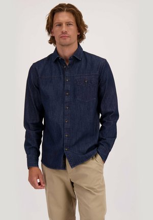 Man in een donker denim hemd met lange mouwen en beige pants, staand met één hand in de zak tegen een effen achtergrond.