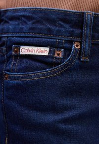 Zilvēkam džinsu bikses ar sarkanu un baltu Calvin Klein etiķeti, vara apdari un kontrastējošu dzeltenu šuvumu gar kabatām.