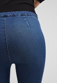 Jeans de denim azul oscuro con un diseño ajustado, detalles en la cinturilla y una textura suave con costuras mínimas visibles.