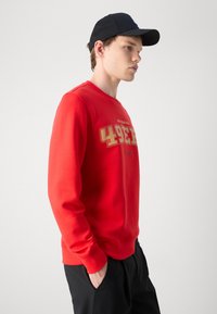 Sweatshirt vermelho com gola redonda, apresentando o texto dourado "San Francisco 49ers". Combinado com um boné preto e calças pretas. Textura suave.