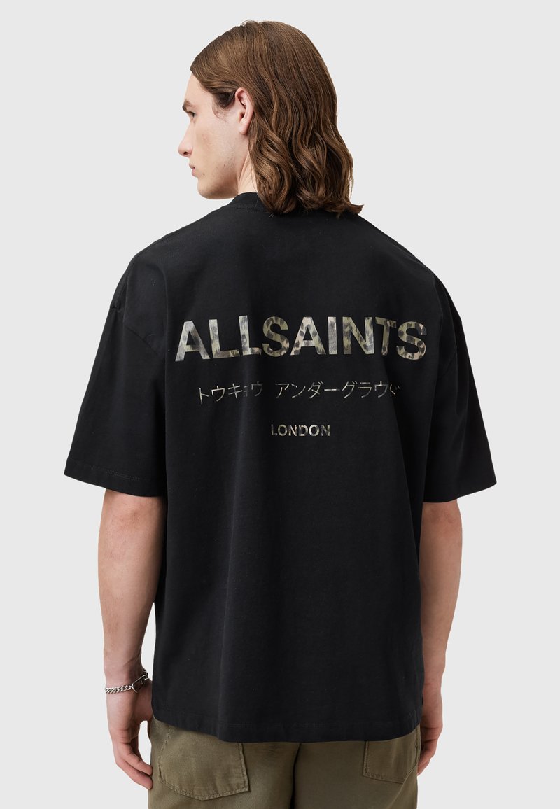 Zwarte katoenen T-shirt met grote camouflage tekst op de achterkant die "ALLSAINTS" leest en kleinere tekst "LONDON" daaronder. Korte mouwen.