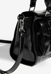 Melnas patentētas ādas rokassomu ar strukturētu formu, kas aprīkota ar sudraba detaļām, regulējamu siksnu un sānu kabatu ar atloka detaļu.